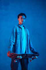 Image showing Caucasian young skateboarder posing on dark neon lighted background