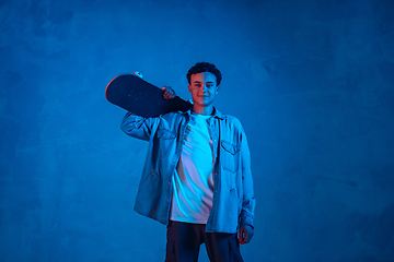 Image showing Caucasian young skateboarder posing on dark neon lighted background