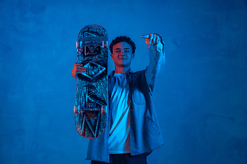 Image showing Caucasian young skateboarder posing on dark neon lighted background