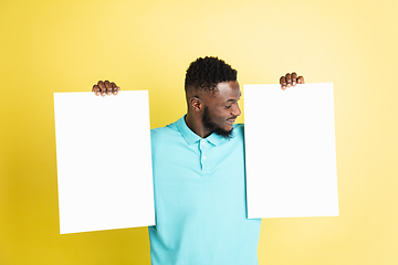 Image showing Young African man with blank sheets of paper isolated over yellow studio background.