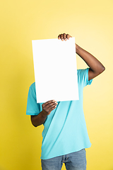 Image showing Young African man with blank sheet of paper isolated over yellow studio background.