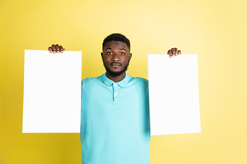 Image showing Young African man with blank sheets of paper isolated over yellow studio background.