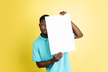 Image showing Young African man with blank sheet of paper isolated over yellow studio background.