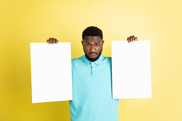 Image showing Young African man with blank sheets of paper isolated over yellow studio background.