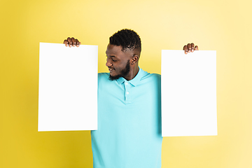 Image showing Young African man with blank sheets of paper isolated over yellow studio background.