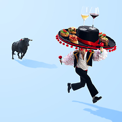 Image showing Traditional corrida, bullfighter in sombrero with tapas and wine glasses is running away from bull on blue background