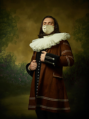 Image showing Young man as a medieval knight on dark background wearing protective mask against coronavirus