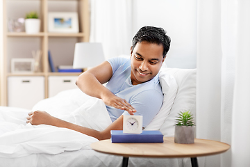 Image showing happy indian man with alarm clock lying in bed