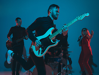 Image showing Young caucasian musicians, band performing in neon light on blue studio background, guitarist in front