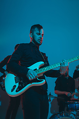 Image showing Young caucasian musicians, band performing in neon light on blue studio background, guitarist in front