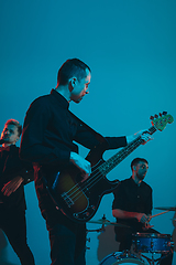 Image showing Young caucasian musicians, band performing in neon light on blue studio background, guitarist in front