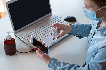 Image showing Young woman in face mask disinfecting gadgets surfaces on her workplace