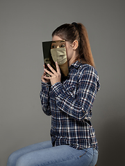 Image showing Be safe and read to become someone else - woman covering face with book in face mask while reading on grey background