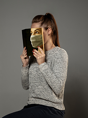 Image showing Be safe and read to become someone else - woman covering face with book in face mask while reading on grey background