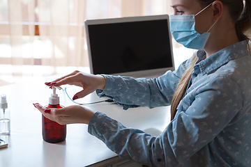 Image showing Young woman in face mask disinfecting gadgets surfaces on her workplace