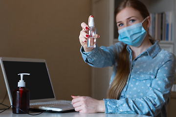 Image showing Young woman in face mask disinfecting gadgets surfaces on her workplace