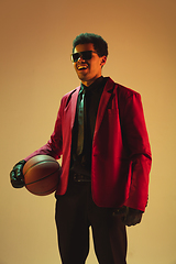 Image showing High-fashion styled man in red jacket playing basketball isolted over brown background