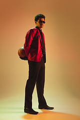 Image showing High-fashion styled man in red jacket playing basketball isolted over brown background