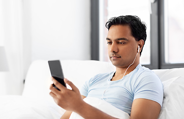 Image showing indian man in earphones with phone in bed at home