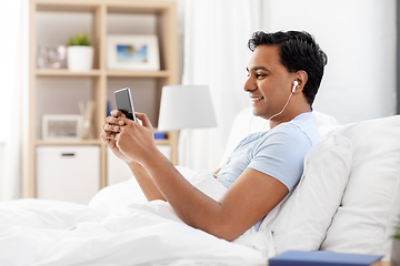 Image showing indian man in earphones with phone in bed at home