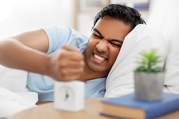 Image showing angry indian man with alarm clock lying in bed