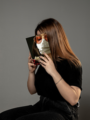 Image showing Be safe and read to become someone else - woman covering face with book in face mask while reading on grey background