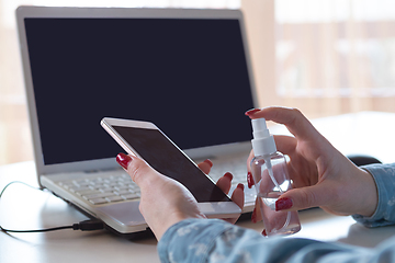 Image showing Young woman in face mask disinfecting gadgets surfaces on her workplace