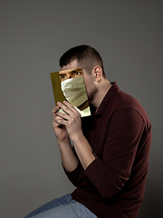 Image showing Be safe and read to become someone else - man covering face with book in face mask while reading on grey background