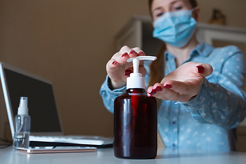 Image showing Young woman in face mask disinfecting gadgets surfaces on her workplace