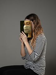Image showing Be safe and read to become someone else - woman covering face with book in face mask while reading on grey background