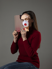 Image showing Be safe and read to become someone else - woman covering face with book in face mask while reading on grey background