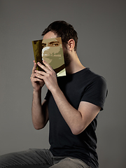 Image showing Be safe and read to become someone else - man covering face with book in face mask while reading on grey background