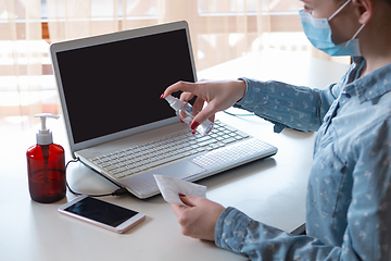 Image showing Young woman in face mask disinfecting gadgets surfaces on her workplace