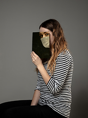 Image showing Be safe and read to become someone else - woman covering face with book in face mask while reading on grey background