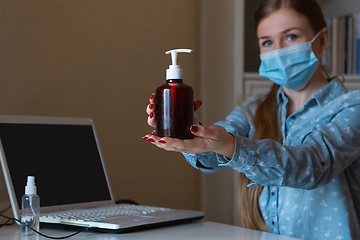 Image showing Young woman in face mask disinfecting gadgets surfaces on her workplace