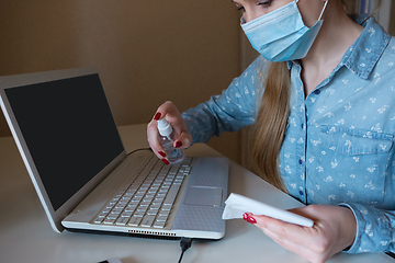 Image showing Young woman in face mask disinfecting gadgets surfaces on her workplace