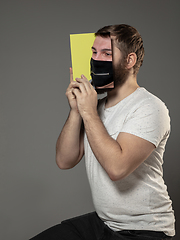 Image showing Be safe and read to become someone else - man covering face with book in face mask while reading on grey background