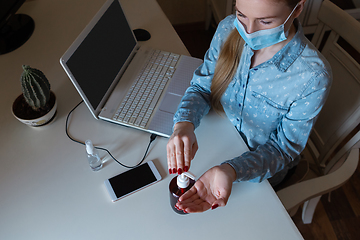 Image showing Young woman in face mask disinfecting gadgets surfaces on her workplace