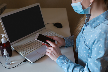 Image showing Young woman in face mask disinfecting gadgets surfaces on her workplace