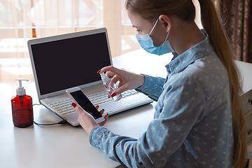 Image showing Young woman in face mask disinfecting gadgets surfaces on her workplace