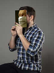 Image showing Be safe and read to become someone else - man covering face with book in face mask while reading on grey background