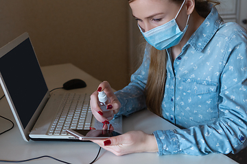 Image showing Young woman in face mask disinfecting gadgets surfaces on her workplace