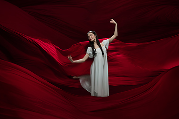 Image showing Young and graceful ballet dancer on billowing red cloth background in classic action. Art, motion, action, flexibility, inspiration concept.