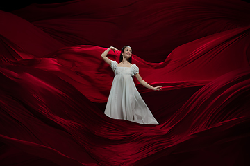 Image showing Young and graceful ballet dancer on billowing red cloth background in classic action. Art, motion, action, flexibility, inspiration concept.