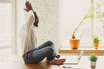 Image showing Young african-american man doing yoga at home while being quarantine and freelance working