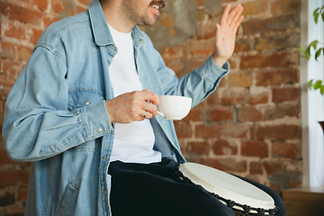 Image showing Caucasian musician playing hand drum during online concert at home isolated and quarantined, greeting band and coffee drinking