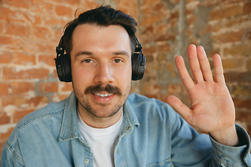 Image showing Caucasian musician in headphones greeting audience before online concert at home isolated and quarantined, cheerful, smiling