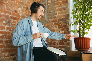 Image showing Caucasian musician playing hand drum during online concert at home isolated and quarantined, greeting band and coffee drinking