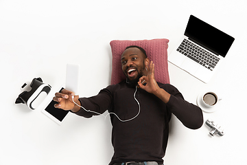 Image showing Emotional african-american man using phone surrounded by gadgets isolated on white studio background, technologies connecting people. Online meeting, selfie