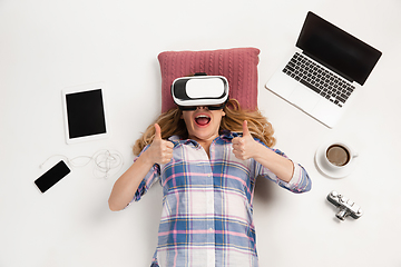 Image showing Emotional caucasian woman using VR-headsed, surrounded by gadgets isolated on white studio background, technologies. Happy shows thumbs up